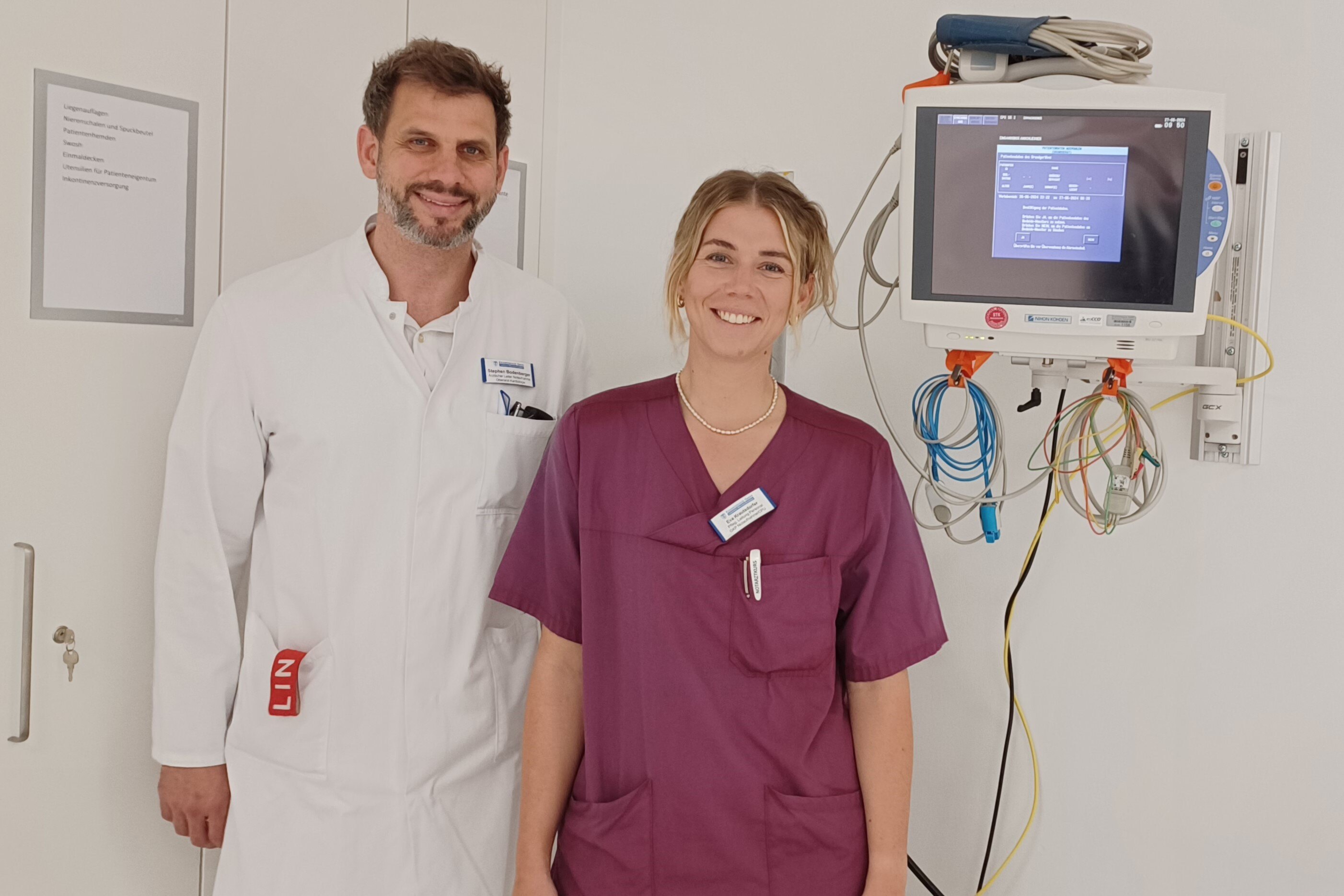 Arzt im weißen Kittel und Pflegekraft in Bordeaux-Kleidung stehen vor einem Monitor im Krankenhaus.