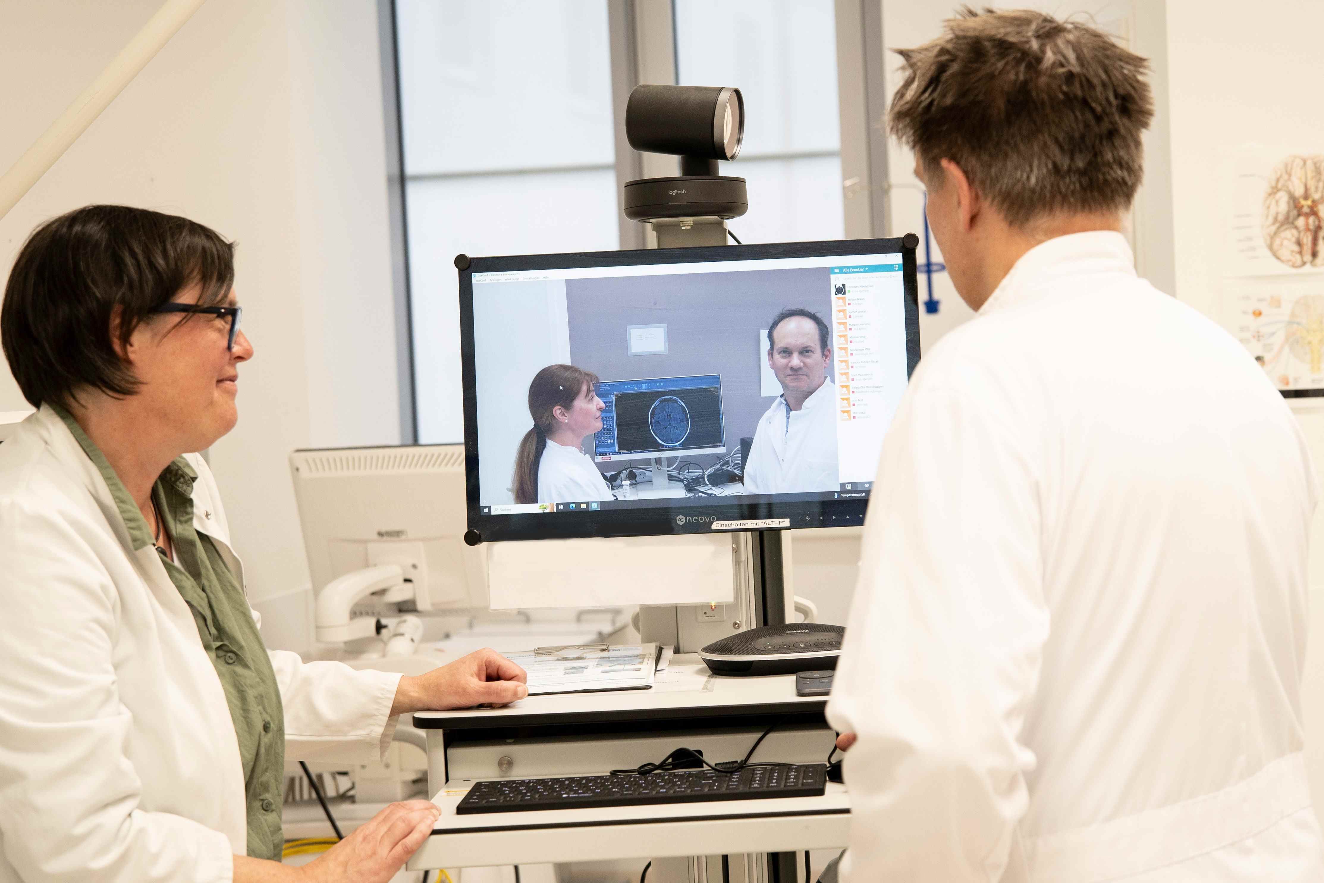 Zwei Ärzte stehen vor einem Monitor und nehmen an einer Videokonferenz mit Kollegen teil