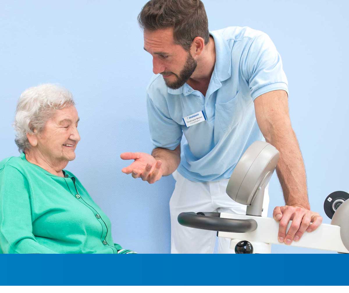 Therapeut spricht freundlich mit einer Seniorin und steht dabei neben einem Trainingsgerät