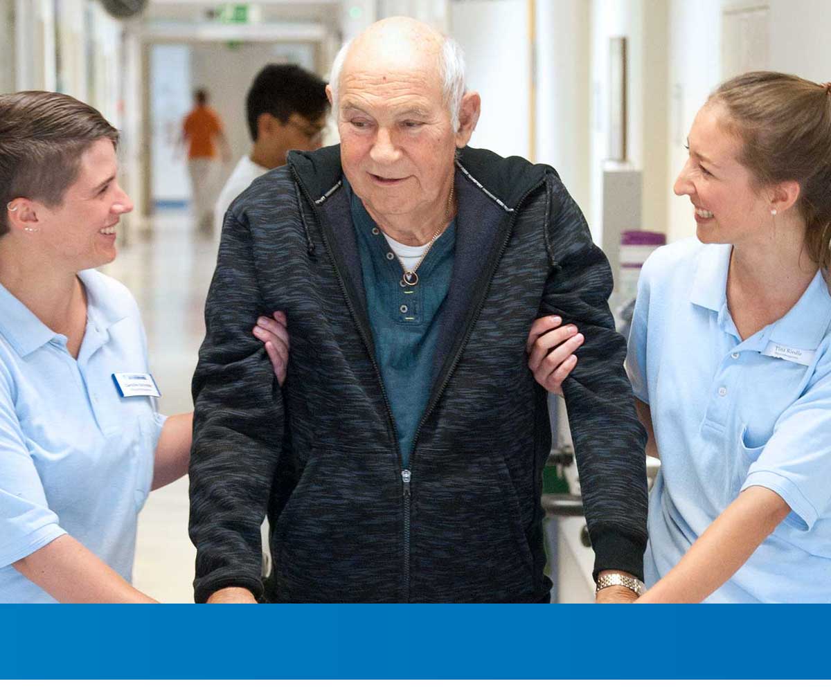 Zwei Pflegekräfte unterstützen einen älteren Mann beim Gehen durch einen Klinikflur.