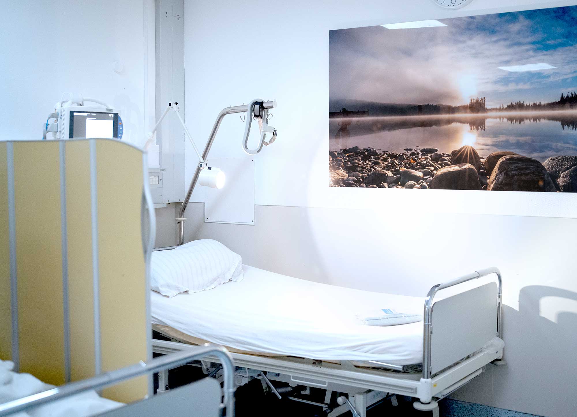 Krankenhauszimmer mit leerem Bett und Landschaftsbild an der Wand.